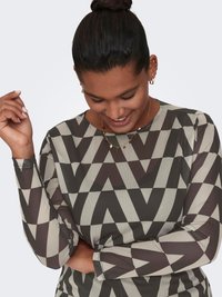 Woman with dark hair in a bun wearing geometric-patterned long-sleeve top, smiling and looking down with one arm crossed and one hand raised.