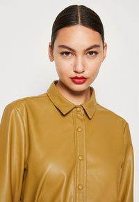 Camicia in ecopelle giallo senape con colletto e chiusura a bottoni a pressione, caratterizzata da una superficie liscia e lucida.