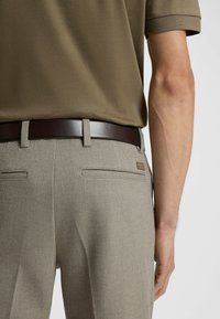 Beige visgraat broek met dubbele achterzakken, gecombineerd met een bruine leren riem. Olijfgroen poloshirt gezien van achteren.