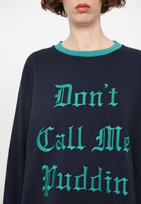 Marineblå oversized hettegenser med turkise detaljer og brodert tekst: "Ikke kall meg Puddin" i fet, stilisert skrift.