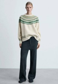 Kremasti pleteni pulover s zelenim i bež geometrijskim uzorcima na ramenu, uparen s teksturiranim tamno sivim širokim hlačama.