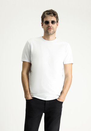 Weißes Kurzarm-T-Shirt aus glattem Stoff, kombiniert mit schwarzen Jeans. Die Accessoires beinhalten runde, getönte Sonnenbrillen. Das Modell steht mit den Händen in den Taschen.
