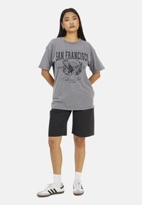 T-shirt oversize gris avec impression graphique noire affichant "SAN FRANCISCO," associé à un short en denim noir et des chaussettes blanches avec des baskets grises.