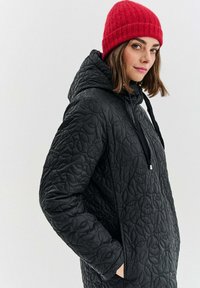 Žena v černé prošívané kapuciované bundě a červené pletené čepici se dívá stranou s rukama v kapsách na jednobarevném pozadí.