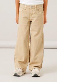 Beige levele trousers made of cotton twill, with front pockets, a button closure, and subtle stitching details. Paired with light-colored sneakers.