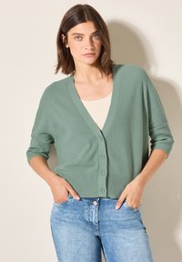Lichtgroene gebreide cardigan met een V-hals, knoopsluiting en driekwartmouwen, gecombineerd met blauwe jeans. Zachte textuur en relaxte pasvorm.