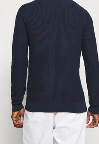 Mężczyzna w granatowym, prążkowanym swetrze z długim rękawem i białych spodniach, pokazany z tyłu na tle jasnej, gładkiej ściany.