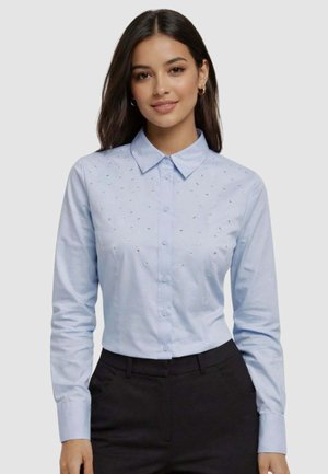 Giovane donna con capelli scuri che indossa una camicia azzurra chiara con piccoli ornamenti sul petto e pantaloni neri, in piedi davanti a uno sfondo semplice.