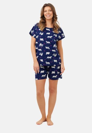 Kobieta w granatowej bluzce z krótkim rękawem i szortach z wzorem białych kotów i myszy, stojąca boso i uśmiechająca się na tle jednolitego tła.