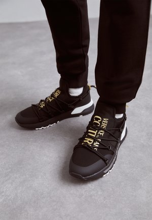 Person iført sorte Versace Jeans Couture sneakers med guldbogstaver, sorte bukser og hvide sokker på en grå gulv.