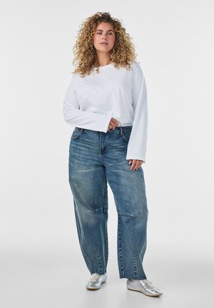 Kvinna med lockigt hår som bär en vit långärmad skjorta, lösa blå jeans och silverfärgade sneakers, stående mot en enfärgad bakgrund.