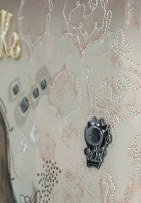 Pelle texturizzata rosa chiaro con motivi di cucitura argentata, borchie decorative e una spilla a forma di gatto nero con una tiara e strass.