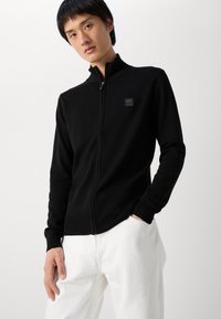 Black zip-up jacket with high collar, ribbed cuffs, and a small brand logo patch on the chest; smooth texture and fitted design.