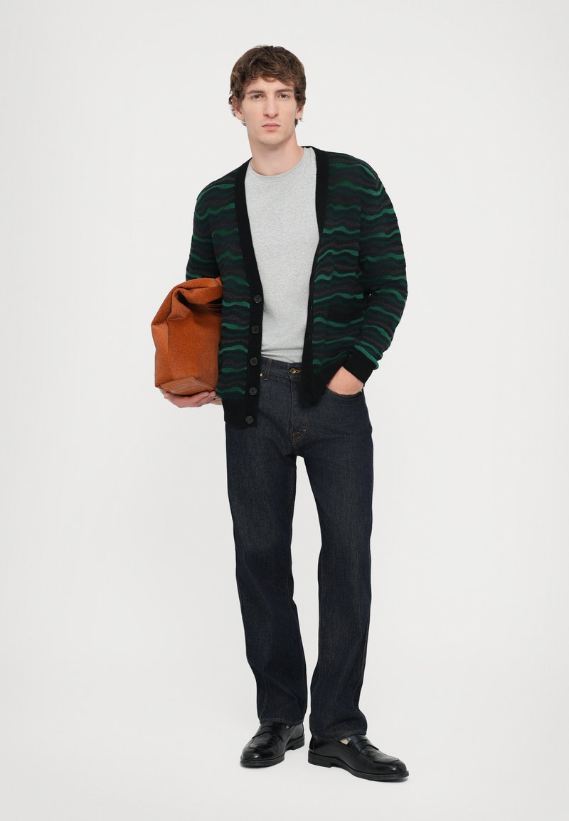 Knitted cardigan with dark green wave pattern, black trim, gray t-shirt, dark jeans, and black shoes; holding a brown bag.
