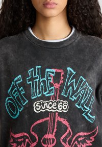 Sudadera negra con textura desgastada, estampado gráfico en azul y rosa que dice "OFF THE WALL desde 66," rodeado de un diseño decorativo.