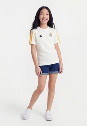 Fille souriante aux longs cheveux noirs portant un maillot de football Real Madrid blanc avec des rayures jaunes, un short en jean et des baskets blanches, debout.