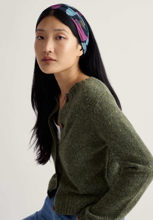 Zelený pletený cardigan s kulatým výstřihem a dvěma hnědými knoflíky. Nošený přes bílý top, doplněný barevnou vzorovanou čelenkou.