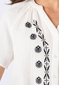 Witte button-up shirt van textuurstof, met zwarte bloemenborduursels langs de sluiting en korte mouwen.