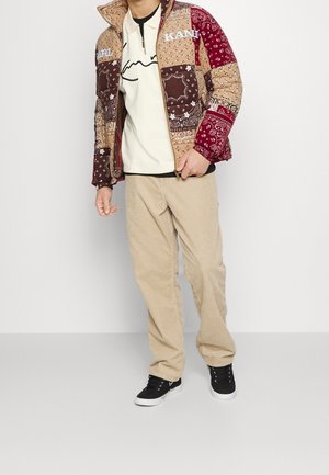 Patchwork jack in bruine en rode tinten met bandana-patronen; crèmekleurig overhemd met zwarte lijntekening; beige ribfluwelen broek; zwarte sneakers.