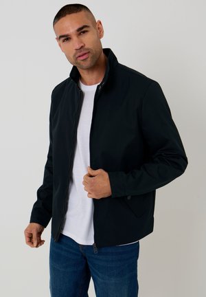 Mand iført en sort lynlåstrøje over en hvid t-shirt og blå jeans, vendt mod fronten med en hånd, der justerer jakkelåsen.