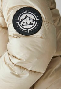 Veste matelassée beige avec un patch circulaire noir et blanc Clarks montrant des montagnes, des points de boussole, et le texte "L'aventure commence ici."
