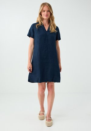 Femme blonde debout portant une robe bleu marine à manches courtes et des chaussures plates en tissu beige sur un fond clair uni.