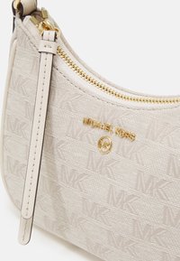 Jasno beżowa torebka wykonana z tkaniny z wytłoczonym wzorem logo MK, złotym suwadłem i skórzanym detalem na pasku. Posiada tabliczkę z nazwą marki.