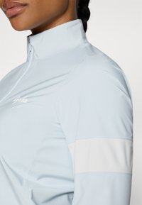 Light blue athletic jacket with a high collar, white shoulder stripe, zip closure, and textured fabric. Features logo on the chest.