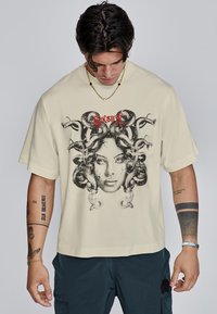 Crème kleurig oversized t-shirt met een grafiek van een vrouwen gezicht omringd door slangen, met daarboven rode tekst. Korte mouwen, relaxte pasvorm.