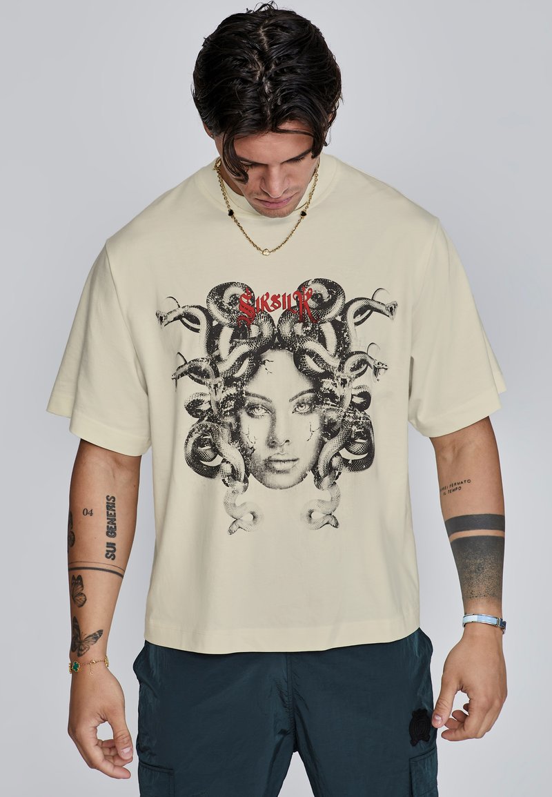 Crème kleurig oversized t-shirt met een grafiek van een vrouwen gezicht omringd door slangen, met daarboven rode tekst. Korte mouwen, relaxte pasvorm.