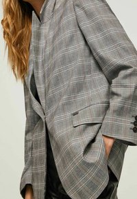 Blazer de cuadros grises con cuello con muescas, corte oversize y acentos de botones negros. Fabricado en un tejido texturizado que presenta líneas blancas y marrones.