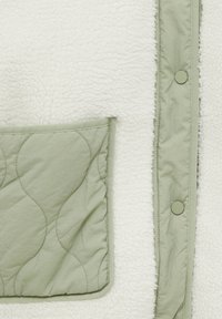 Poche matelassée vert clair et patte pressionnée sur tissu polaire blanc texturé doux d'un vêtement.
