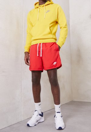 Mężczyzna ubrany w żółtą bluzę z kapturem, czerwone spodenki Nike z białym sznurkiem, białe skarpetki Nike oraz białe buty sportowe z niebieskimi akcentami, stojący przy jednolitej ścianie.