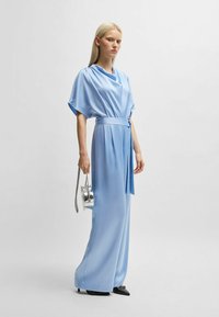 Lichtblauwe satijnen jumpsuit met een gedrapeerde halslijn, wijdvallende broekspijpen en een gestrikte taille. Gestyled met een kleine zilveren handtas en zwarte hakken.