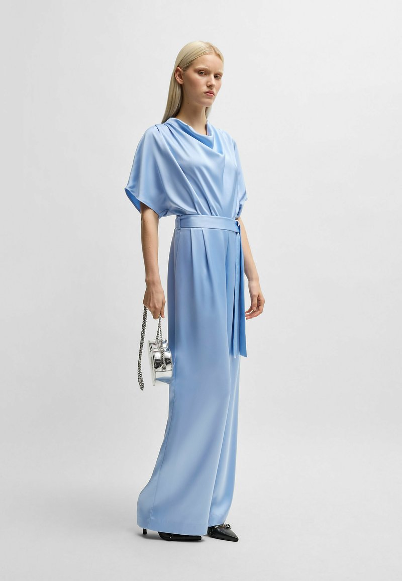 Lichtblauwe satijnen jumpsuit met een gedrapeerde halslijn, wijdvallende broekspijpen en een gestrikte taille. Gestyled met een kleine zilveren handtas en zwarte hakken.