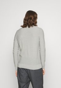 Person med axellångt brunt hår som bär en ljusgrå långärmad tröja och mörkgrå byxor, sedd bakifrån.