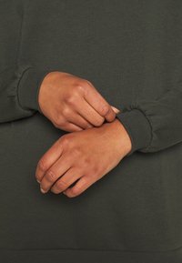 Ciemnozielona bluza z marszczonymi mankietami, pokazana z rękami złączonymi na nadgarstkach. Materiał wydaje się miękki o gładkiej fakturze.