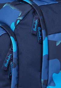 Blauer Rucksack mit dunklen Marine- und Cyan-Camouflage-Mustern. Verfügt über Reißverschlusszüge mit dem Markennamen "Satch" in Blau, aus einem strukturierten Stoff.