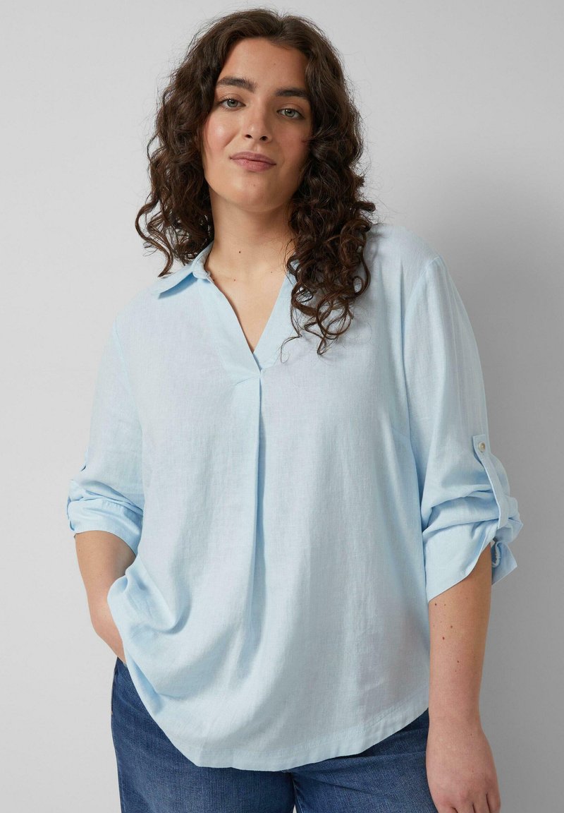 Femme aux cheveux bouclés portant une chemise à manches longues bleu clair avec les manches retroussées et un jean bleu, debout devant un fond uni.