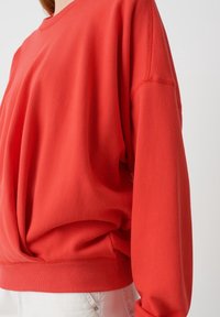 Sudadera roja de gran tamaño hecha de tela suave, con un cuello redondo, hombros caídos y un dobladillo ribeteado con sutiles detalles de costura.