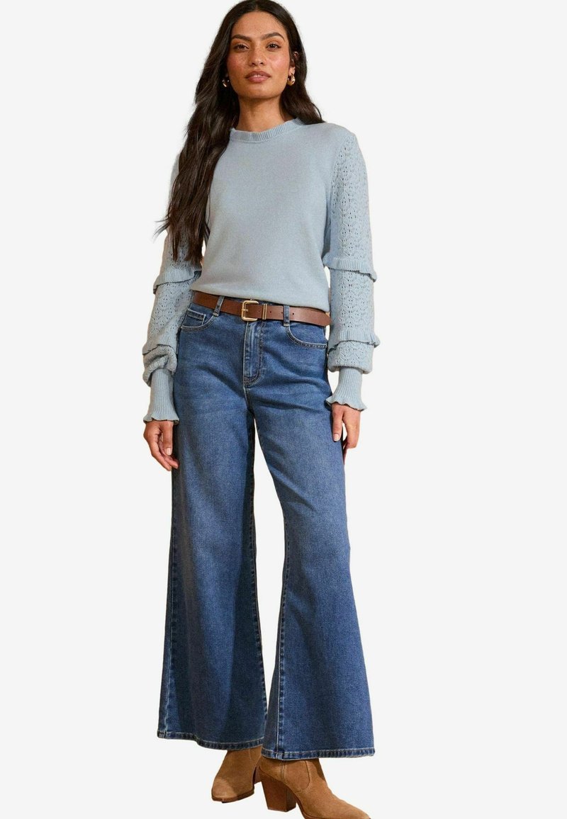 Svetlo modra pletena jopica z teksturiranimi rokavi, v kombinaciji s širokimi modrimi džinsom in svetlim čeveljema do gležnjev. Rjav pas poudarja pas.