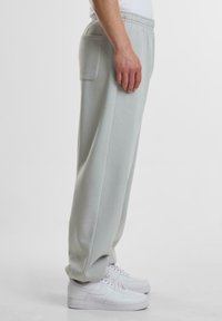 Helle graue Jogginghosen mit einem elastischen Bund, Seitentasche und schmal zulaufenden Beinen, kombiniert mit weißen Sneakers vor einem schlichten Hintergrund.