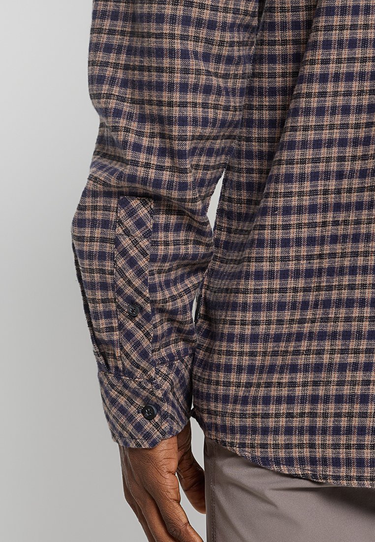 Manche d'une chemise à carreaux bleu et beige avec des boutons noirs, portée par une personne à la peau foncée, présentée à côté d'un pantalon marron clair.