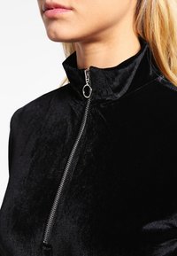 Schwarzes Samt-Zip-up-Oberteil mit Stehkragen, glatter Textur und einem silbernen Reißverschluss mit einem runden Zugband.