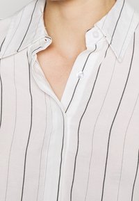 Camicia bianca con sottili righe verticali nere, dotata di una pattina con bottoni e una morbida texture del tessuto.