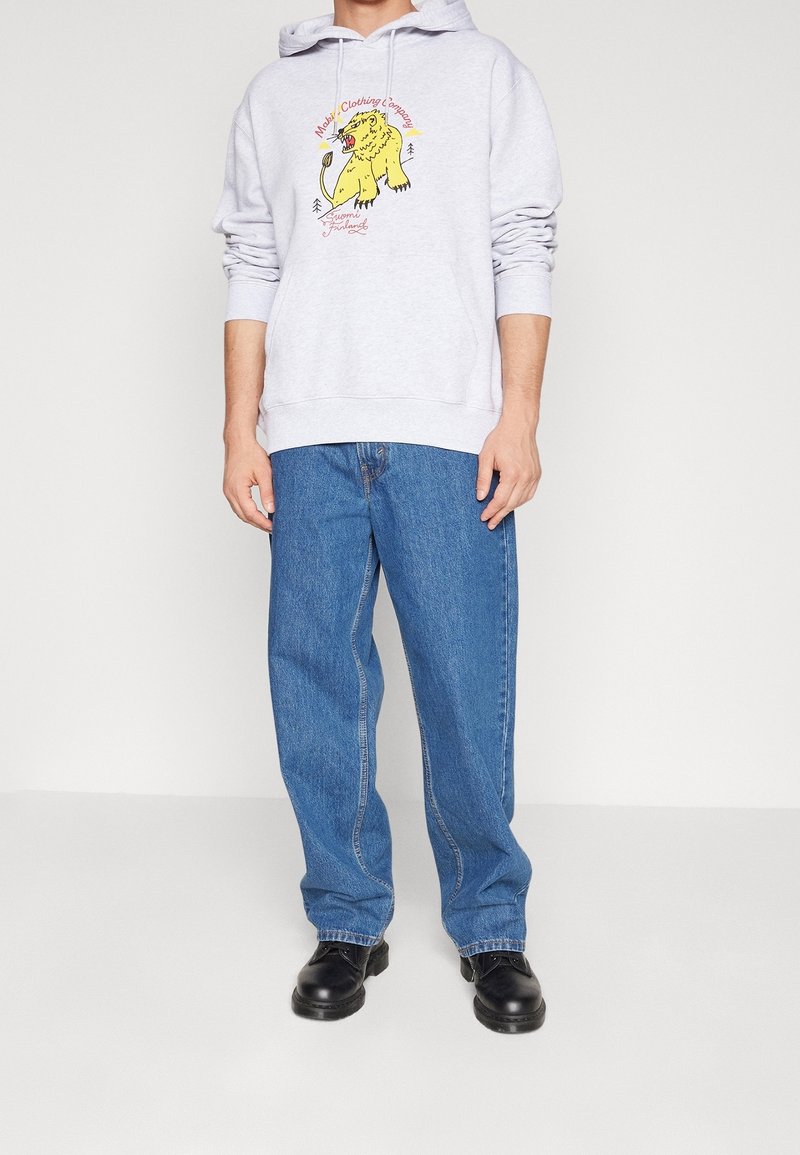 Personne portant un sweat à capuche gris clair avec un motif d'ours jaune et du texte, jean bleu ample et chaussures noires, debout sur fond blanc.