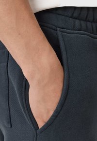 Ciemnoszare dresy z teksturowanej tkaniny i elastycznym pasem, z boczną kieszenią i luźnym krojem, przedstawione z ręką w kieszeni.