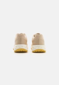 Un par de zapatillas deportivas beige con medias suelas blancas y suelas amarillas, vistas desde la parte posterior sobre un fondo blanco.