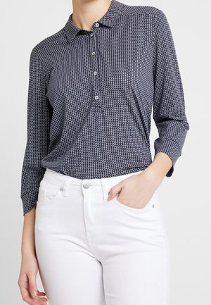 Kobieta nosząca granatową, wzorzystą koszulkę polo z długim rękawem, wpuszczoną w białe, wysokowalne jeansy, z jedną ręką w kieszeni.