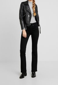 Veste de motard en cuir noir portée sur un pull à côtes gris, associée à un jean noir évasé taille haute et des bottines noires.